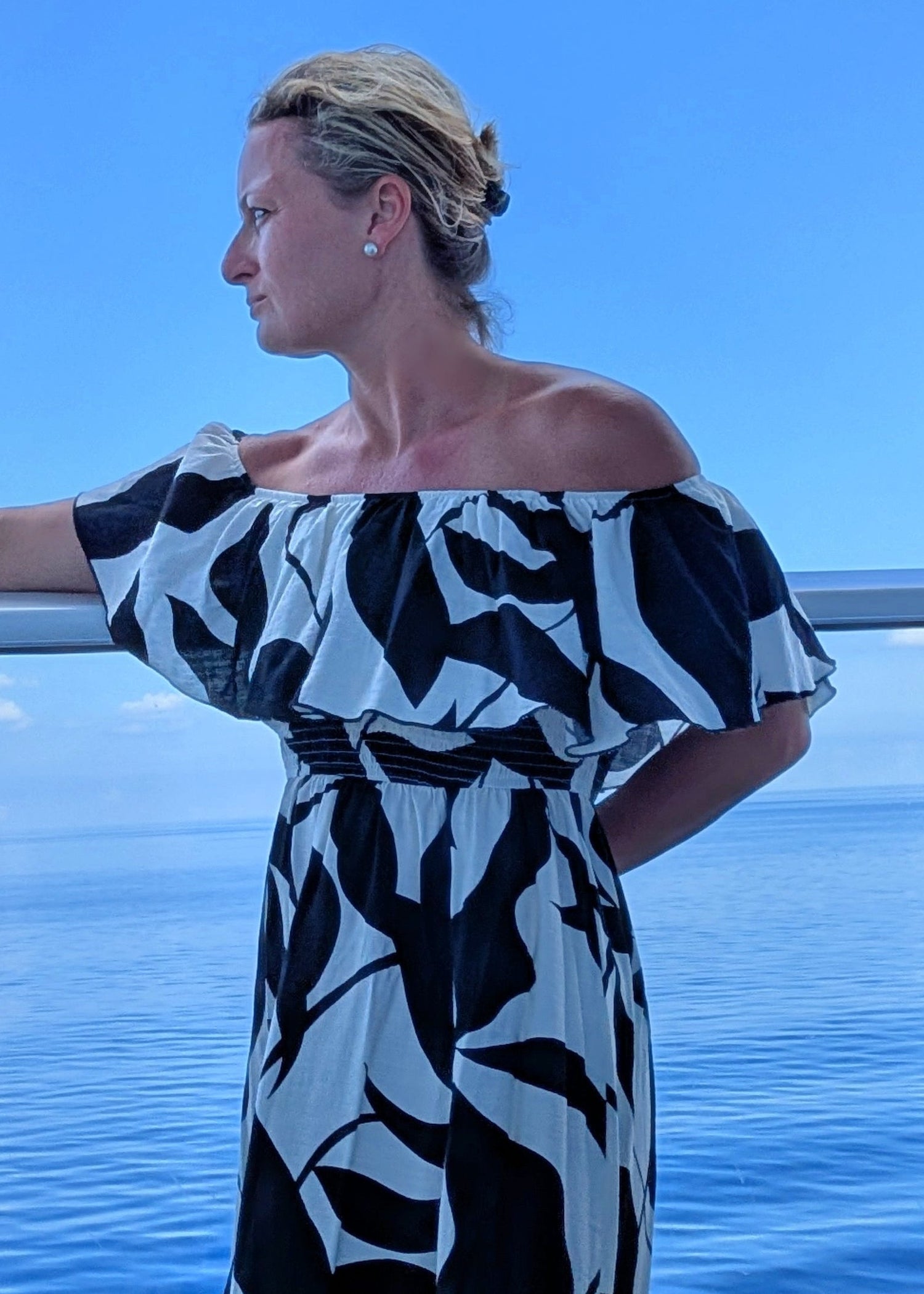 Woman in a black and white patterned dress standing by a railing with a blue sky and water background